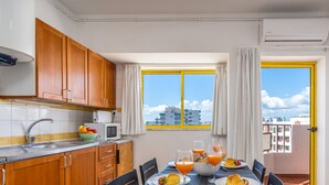A compact kitchen with wooden cabinets, a microwave, and a sink. A dining table with breakfast items is set near a window with yellow frames and curtains. - #303 Atlântida Praia da Rocha by Home Holidays (Portimão)