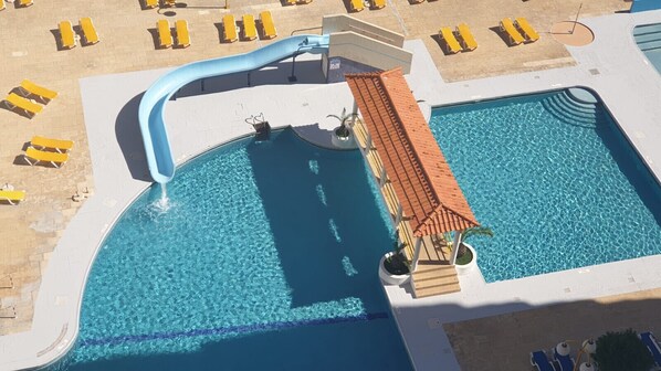 A large outdoor swimming pool with a slide, surrounded by sun loungers and a small pavilion.