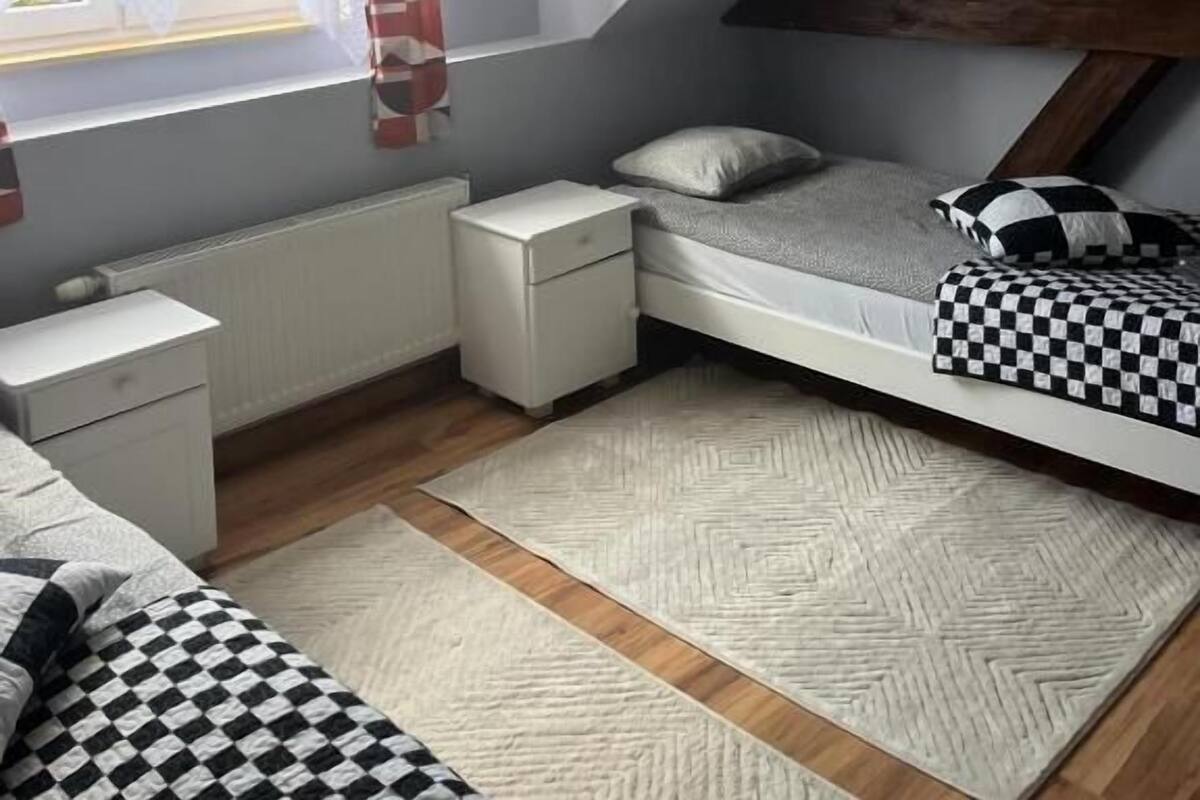 Ein Schlafsaalzimmer mit schrägverdeckter Decke, einem Etagenbett, einem kleinen Nachttisch, einem Heizkörper und einem Fenster mit Vorhängen.