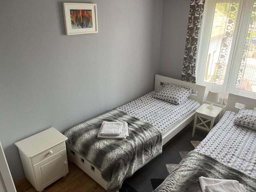 A small, neatly arranged bedroom with two single beds, a bedside table with a lamp, a nightstand, and a framed picture on the wall.