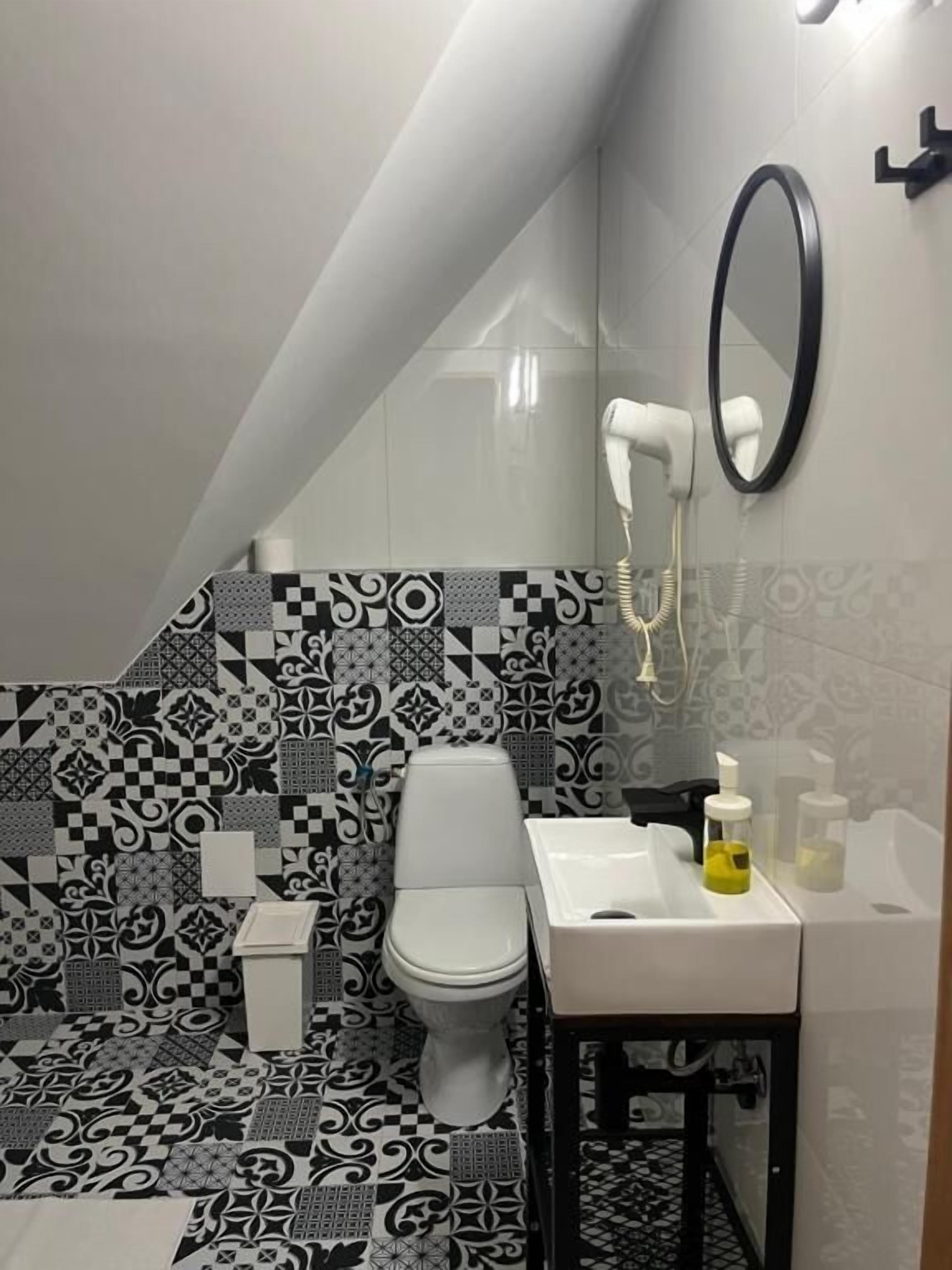 Une salle de bain avec des toilettes blanches, un lavabo et un miroir. Le sol est recouvert de carreaux aux motifs noirs et blancs.