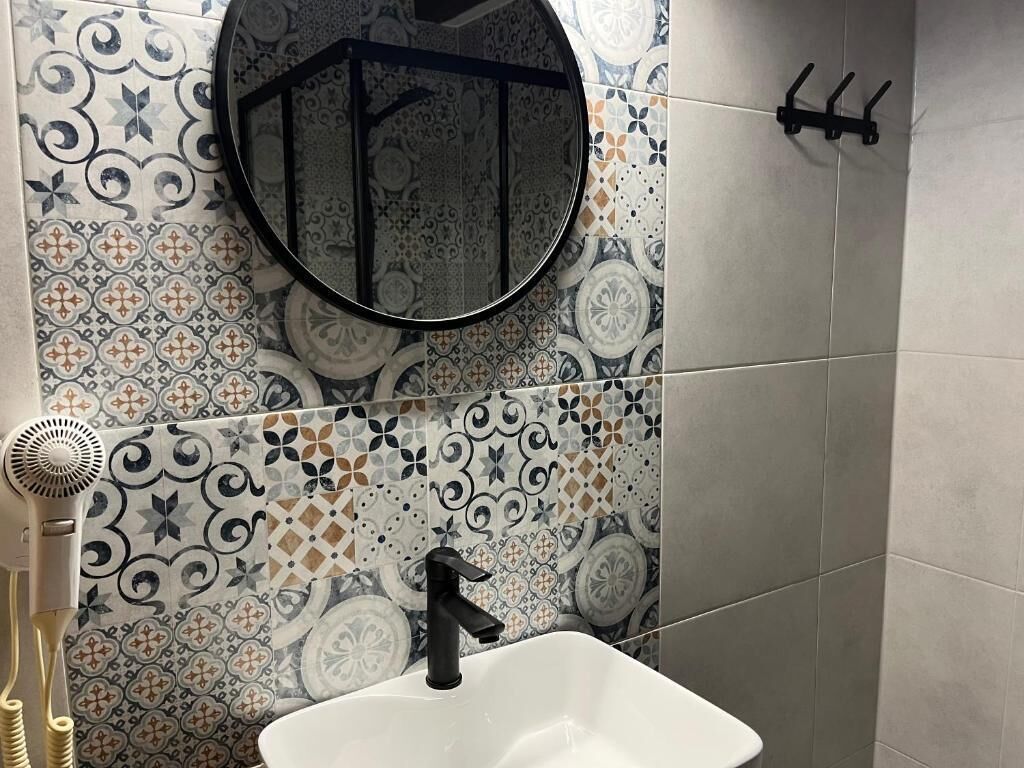 Un bagno con un lavandino bianco, un rubinetto nero e uno specchio rotondo. La parete è decorata con piastrelle a motivi.
