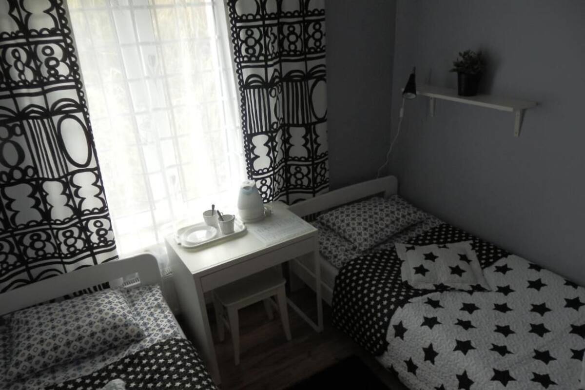 Ein kleiner, schummrig beleuchteter Raum mit zwei Betten, eines mit einer quiltartigen Sternenmusterdecke und das andere mit einem geometrischen Muster. Ein kleiner Tisch mit Tasse und Untertasse, sowie ein Regal mit einer Pflanze.