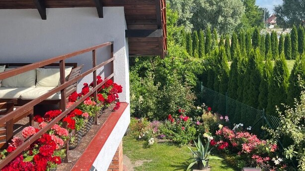 Ein hölzerner Balkon mit weißer Reling, einem Bett und einem Garten mit verschiedenen Blumen und Sträuchern.