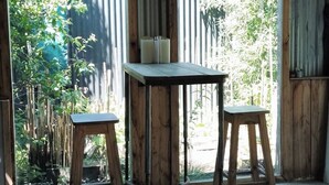 Une pièce avec un sol en béton, deux tabourets en bois, une petite table et une fenêtre à cadre métallique.