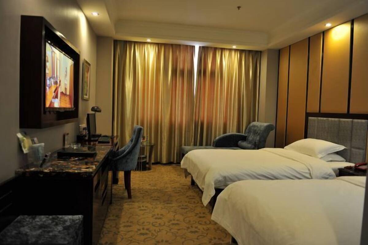 A hotel room with two beds, a desk with a chair, a television, and a large window with curtains.