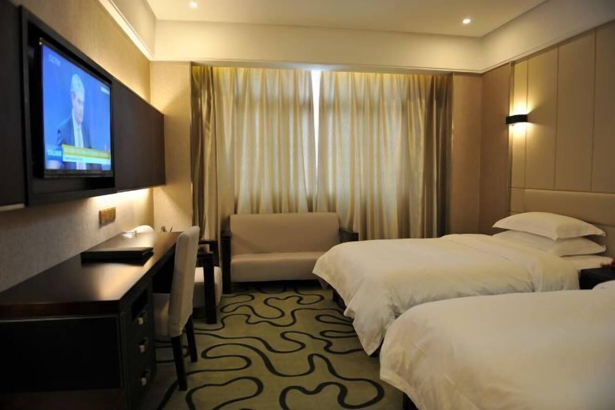 A hotel room with a TV mounted on the wall, a desk, a sofa, and two beds.