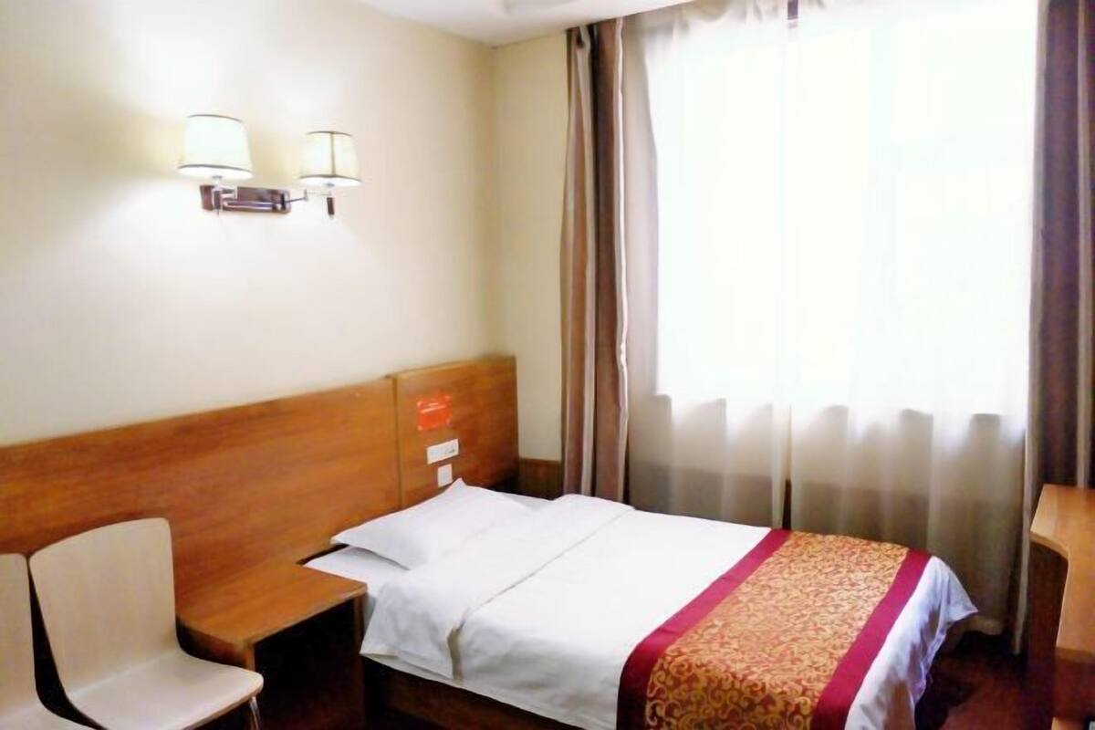 Habitación de hotel con cabecera de madera, cama con ropa de cama blanca y una colcha roja y dorada, mesita de noche, una silla y una ventana con cortinas.