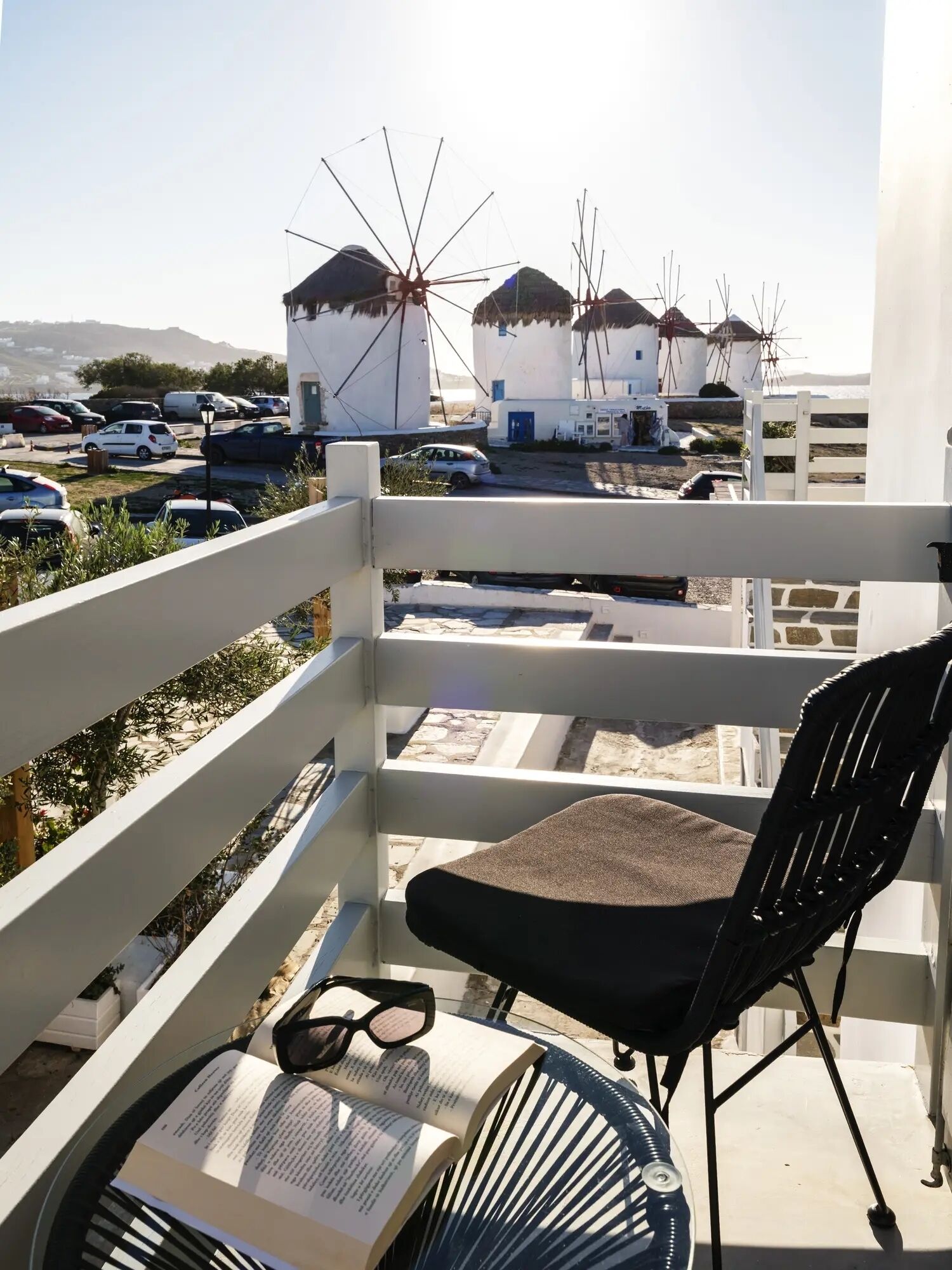 A balcony with a chair, a pair of glasses, and an open book. In the background, there are windmills and parked cars.