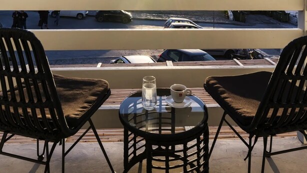 A terrace with two black chairs and a small table, overlooking a coastal area with windmills and a clear sky. - Sunset Windmills Houses (Mykonos)