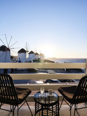 A terrace with two black chairs and a small table, overlooking a coastal area with windmills and a clear sky. - Sunset Windmills Houses (Mykonos)