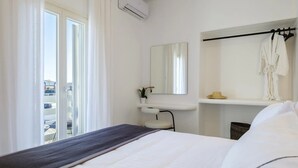 A hotel room with a bed, a bedside table, a mirror, a towel rack, and a window with curtains. - Sunset Windmills Houses (Mykonos)
