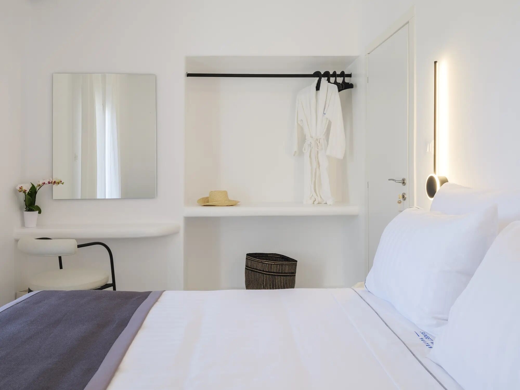 A hotel room with a bed, a bedside table, a mirror, a robe hanging on a rack, and a small trash bin.