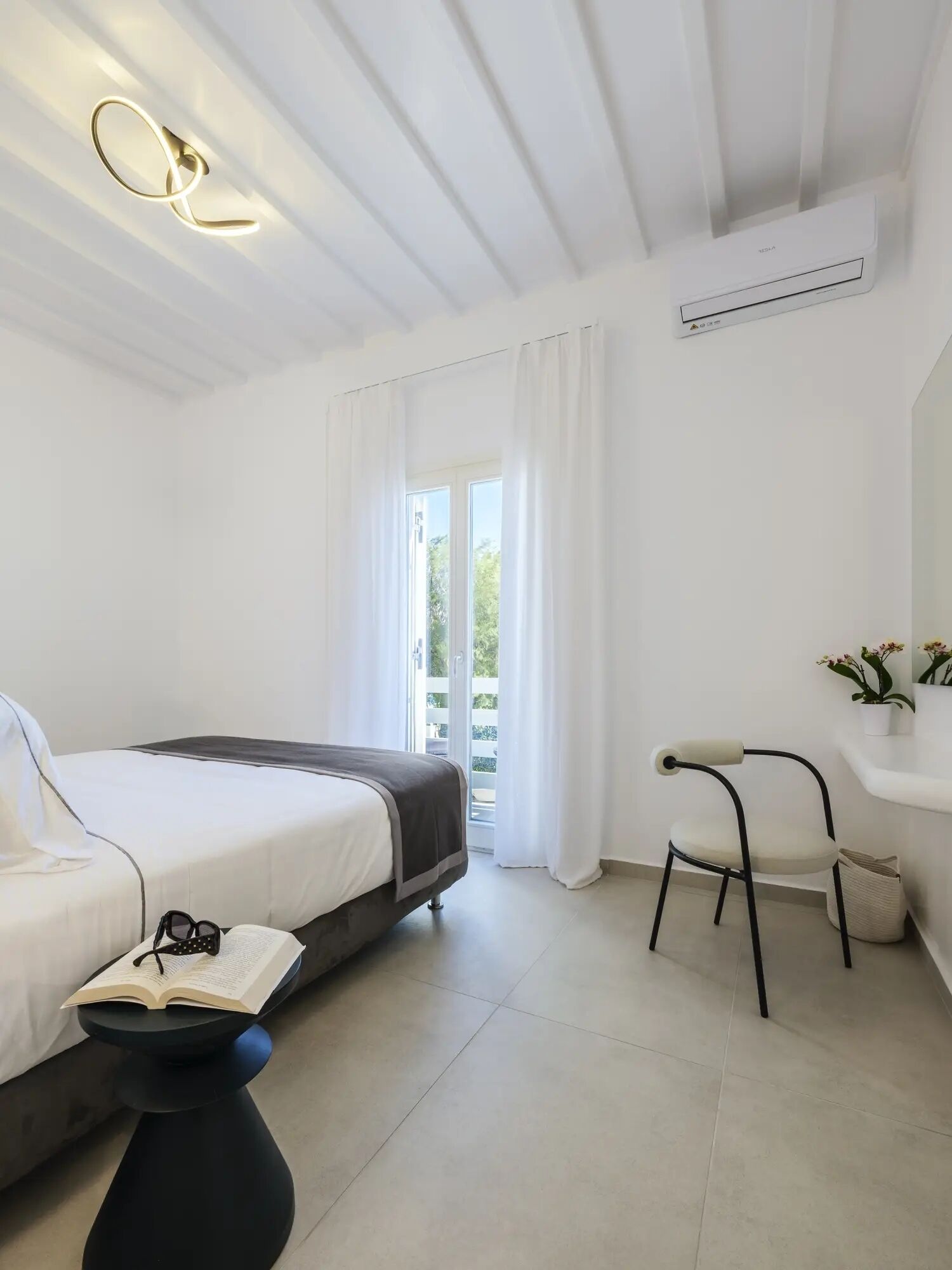 A modern hotel room with a bed, a small side table with a book, a chair, and a window with curtains.