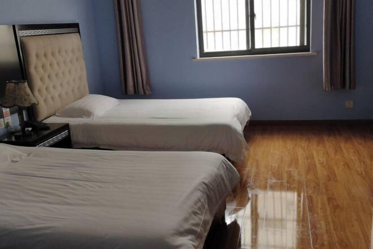 Quarto de hotel com duas camas, uma mesinha de cabeceira, uma luminária e uma janela com cortinas.