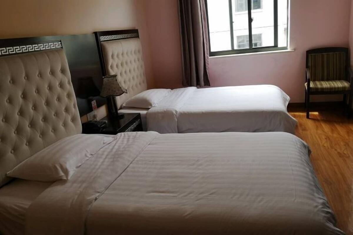 Quarto de hotel com duas camas, mesa de cabeceira, luminária, cadeira e janela com cortinas.