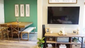 A dining area with a wooden table and chairs, a bench, framed artwork on the wall, and a flat-screen TV mounted above a wooden console with decorative items.