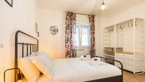 A neatly made bed with a headboard, a bedside lamp, a window with curtains, and a white wardrobe with shelves. - Domus Aqua Alexandrina by Rentbeat (Roma)