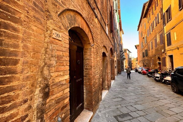 Una stretta strada acciottolata fiancheggiata da edifici storici in mattoni, con un'imponente porta ad arco e una targa con il numero civico.
