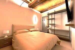 A bedroom with a large bed, a nightstand with a lamp, a wardrobe with shelves, and a ceiling with exposed beams.