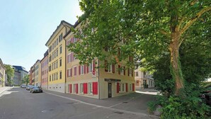 A street view with a row of colorful buildings, a tree-lined sidewalk, and parked cars. - book2stay: modern Apartment at Top Place (Olma Messen) (St. Gallen)