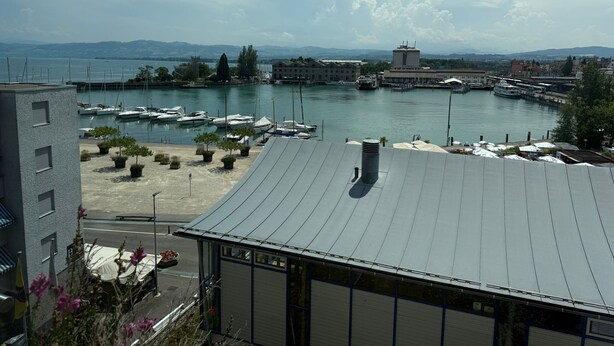 A marina with boats, a building with a curved roof, and a view of distant mountains. - book2stay: Lakefront Apt w/Balcony, near Harbor & Station (Romanshorn)