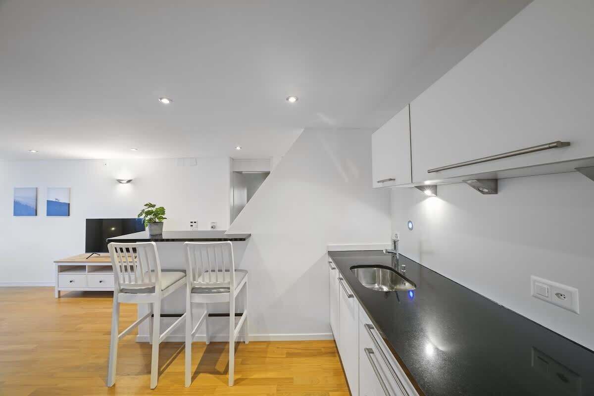 A modern kitchen with white cabinetry, a black countertop, and a stainless steel sink. There are two white chairs and a small table with a plant.