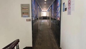 Un pasillo con suelo de madera, paredes de cristal y una lámpara en el techo. Hay letreros en las paredes y un cartel sobre la puerta.