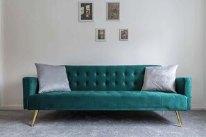 A teal velvet sofa with gold legs, two pillows, and a grey carpeted floor.