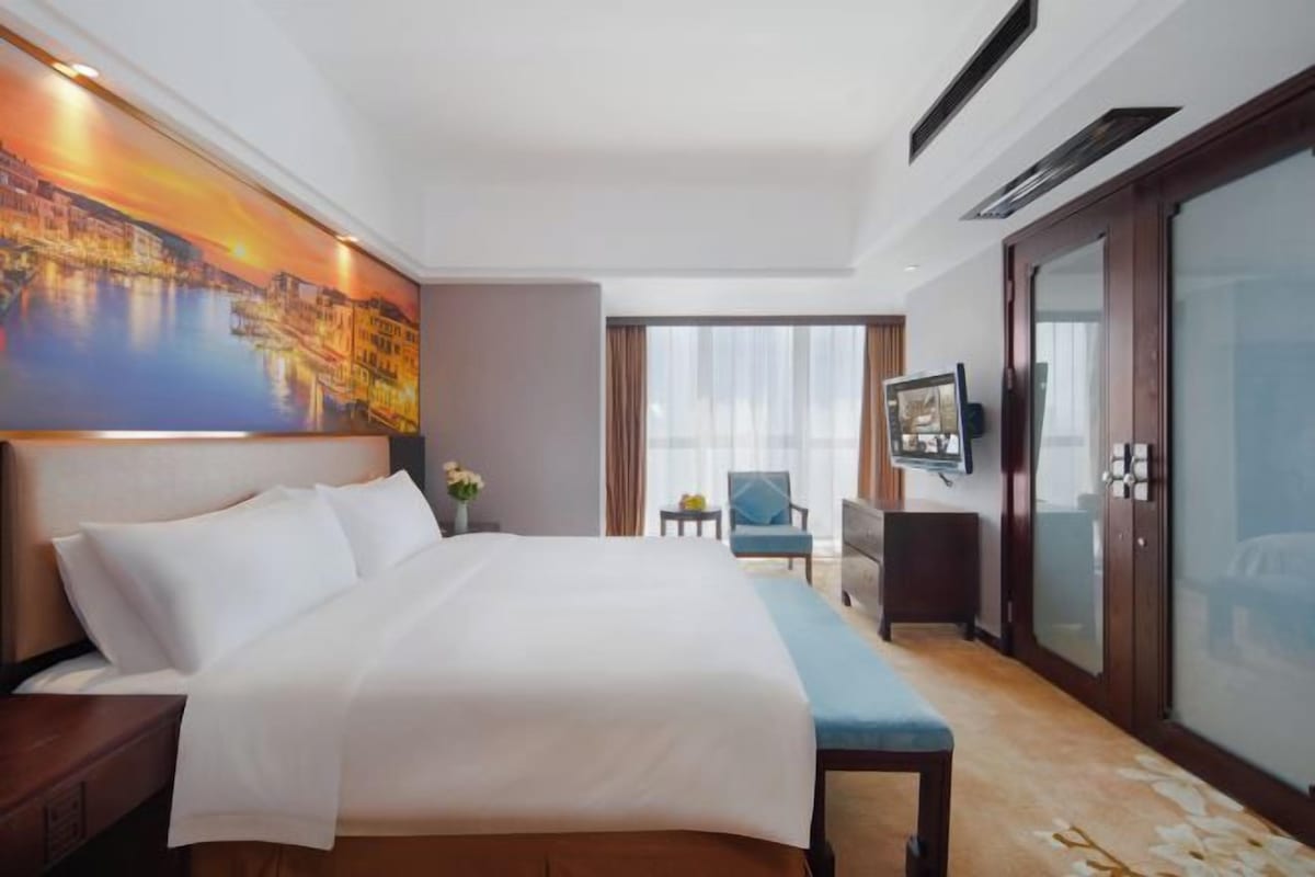 A hotel room with a large bed, a painting on the wall, a TV, and a view of the outdoors.
