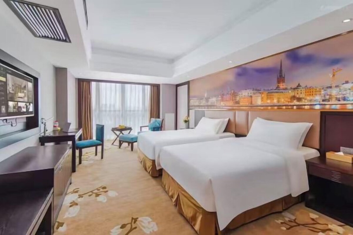 A hotel room with two beds, a desk, a chair, and a large window.