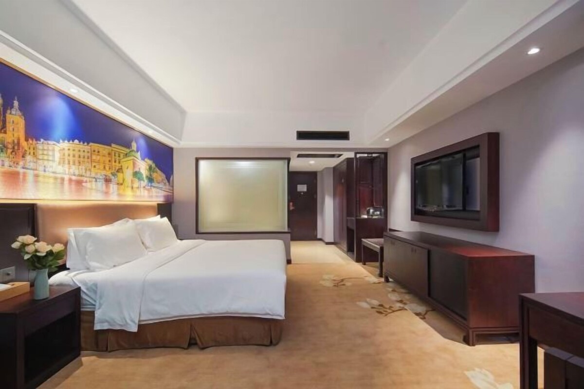 A hotel room with a large bed, a flat-screen TV, a painting on the wall, and a carpeted floor.