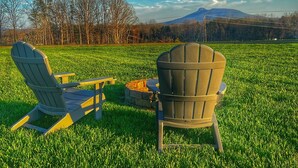 Two Adirondack chairs facing a stone fire pit in a grassy field with a mountain view.