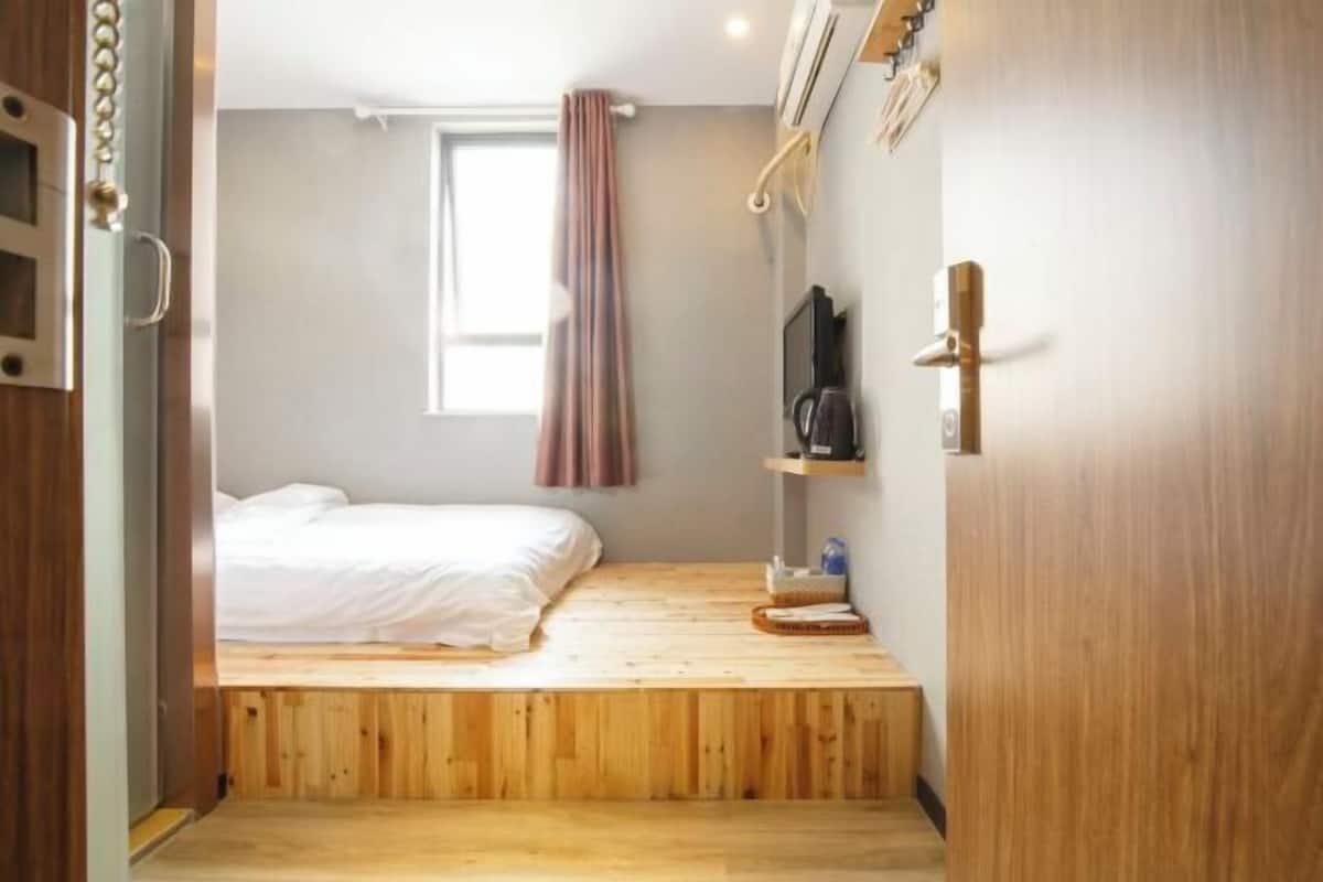 A compact room with a bed, a window with curtains, a shelf with items, and a door.