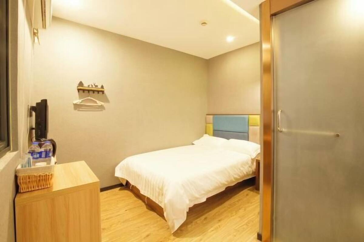 A bedroom with a bed, a wooden desk, a wall-mounted shelf, and a door.