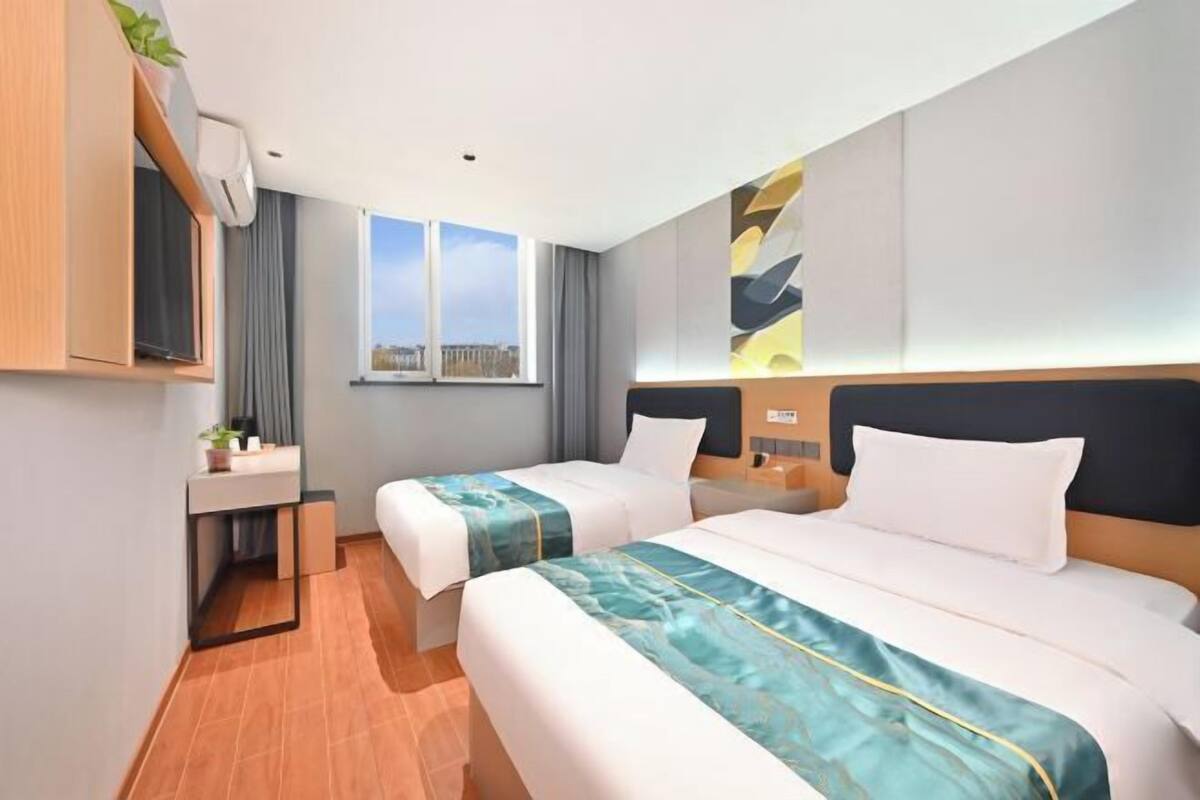 A hotel room with two beds, a desk, a window with a view, and a wall-mounted air conditioner.
