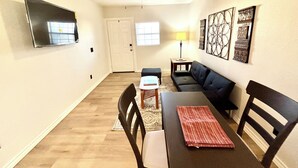A living room with a ceiling fan, a flat-screen TV mounted on the wall, a dark-colored sofa, a dining table with chairs, and a patterned rug. - Casita Del Sol (Big Spring)