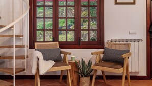 Een kamer met een houten spiltrap, twee houten stoelen met kussens, een tafeltje, een potplant en een raam met uitzicht op groen.