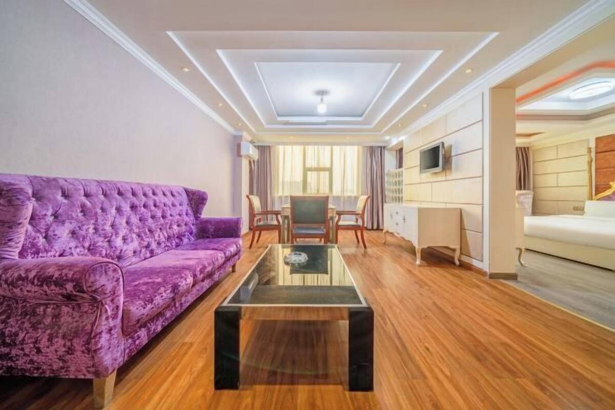 A modern living room with a purple sofa, a glass coffee table, wooden flooring, a TV mounted on the wall, and a ceiling with recessed lighting.
