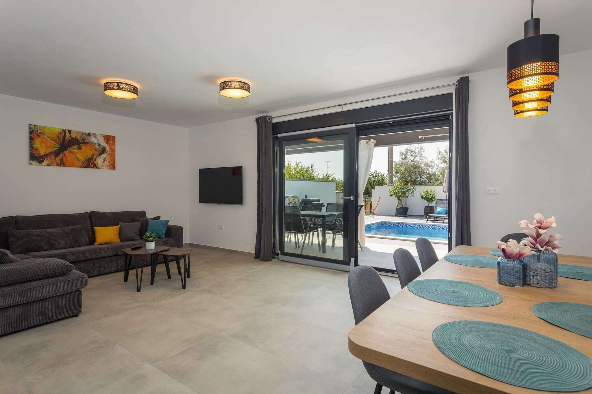 Ein modernes Wohnzimmer mit einer Couch, einem Couchtisch und einem Esstisch mit Platzdeckchen. Ein großes Fenster bietet Blick auf einen Pool und Gartenmöbel.