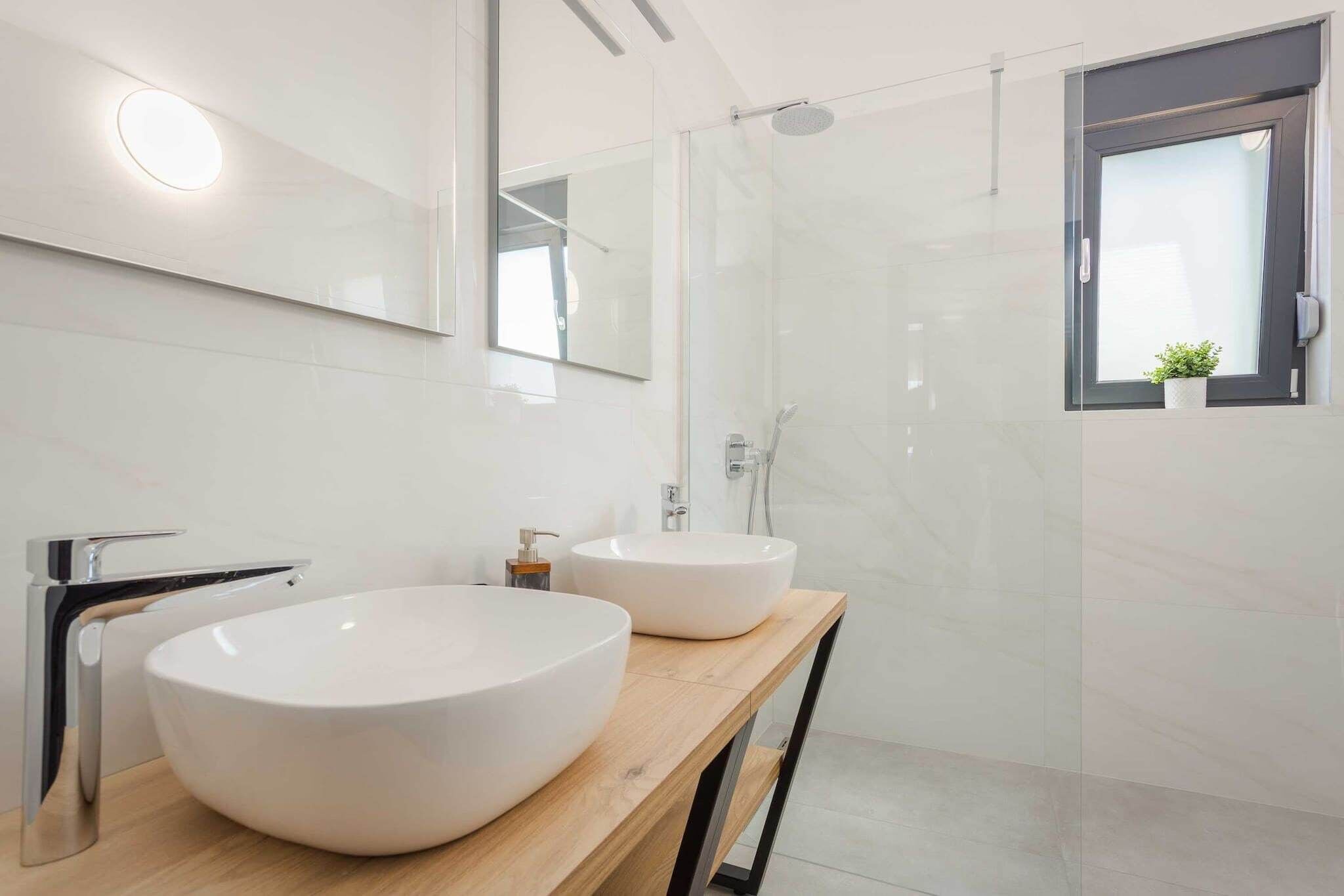 Ein modernes Badezimmer mit zwei großen weißen Waschbecken, einem Holzbadezimmer-Möbelstück, einer Glasdusche und einem kleinen Topfpflanzen auf der Fensterbank.