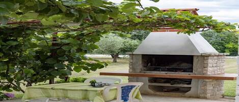 An outdoor grill area with a stone chimney, a table with chairs, and a pergola with green leaves.