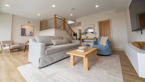 Un soggiorno moderno con un divano grigio, un tavolino in legno e una poltrona blu. Una scala con ringhiere in legno conduce a un piano superiore.