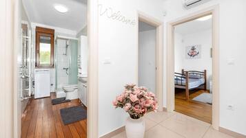A modern bathroom with a shower, toilet, and sink. There is a welcome sign on the wall and a vase with flowers in the hallway.