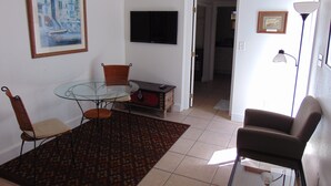 Een woonkamer met een glazen tafel, twee stoelen, een televisie, een klok en een lamp.