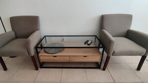 Een kamer met een salontafel met glazen blad, twee fauteuils en een houten bijzettafeltje.