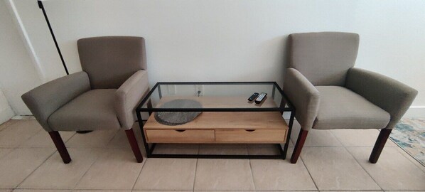 A room with a glass-top coffee table, two armchairs, and a wooden side table.