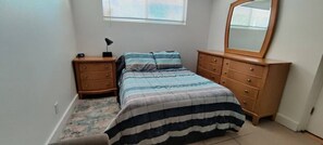 A bedroom with a bed, a nightstand, a dresser, a mirror, and a window with blinds. - Sarasota Springs Apartment (SARASOTA)