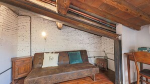 Een rustieke kamer met bakstenen muren, een houten plafond, een bruine leren bank met kussens, een houten stoel en een klein houten kastje.