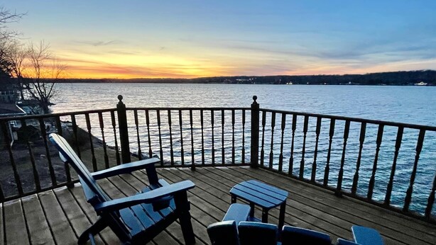 A wooden deck with chairs and a table overlooking a body of water at sunset. - Waterfront Lake House with game room, private pier and kayaks in Lake Geneva (Lake Geneva)